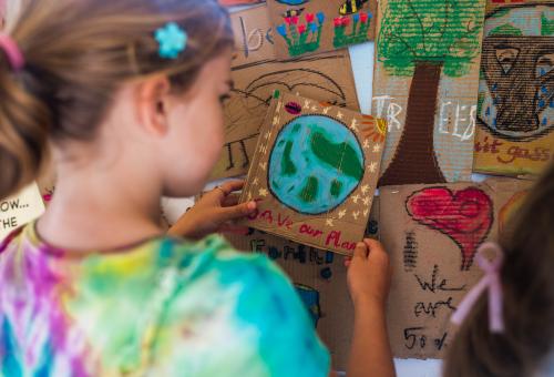 Child placing a handmade cardboard placard with a 'Save our planet' design onto a wall of placards
